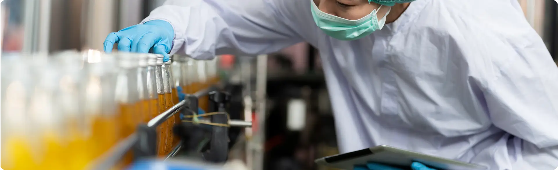 A quality control inspector wearing a hairnet, face mask, and blue gloves, touching or checking glass bottles filled with an amber or orange liquid while holding a digital tablet.